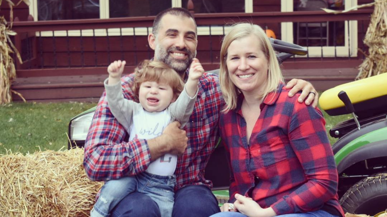 Smiling mom and dad, both wearing blue and red plain flannel tops, with their happy baby boy with his arms in the air.