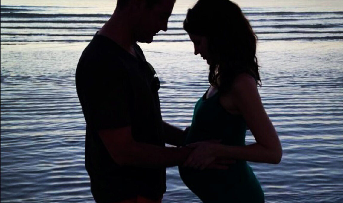 Silhouettes of dad and pregnant mom standing on the beach at sunset.