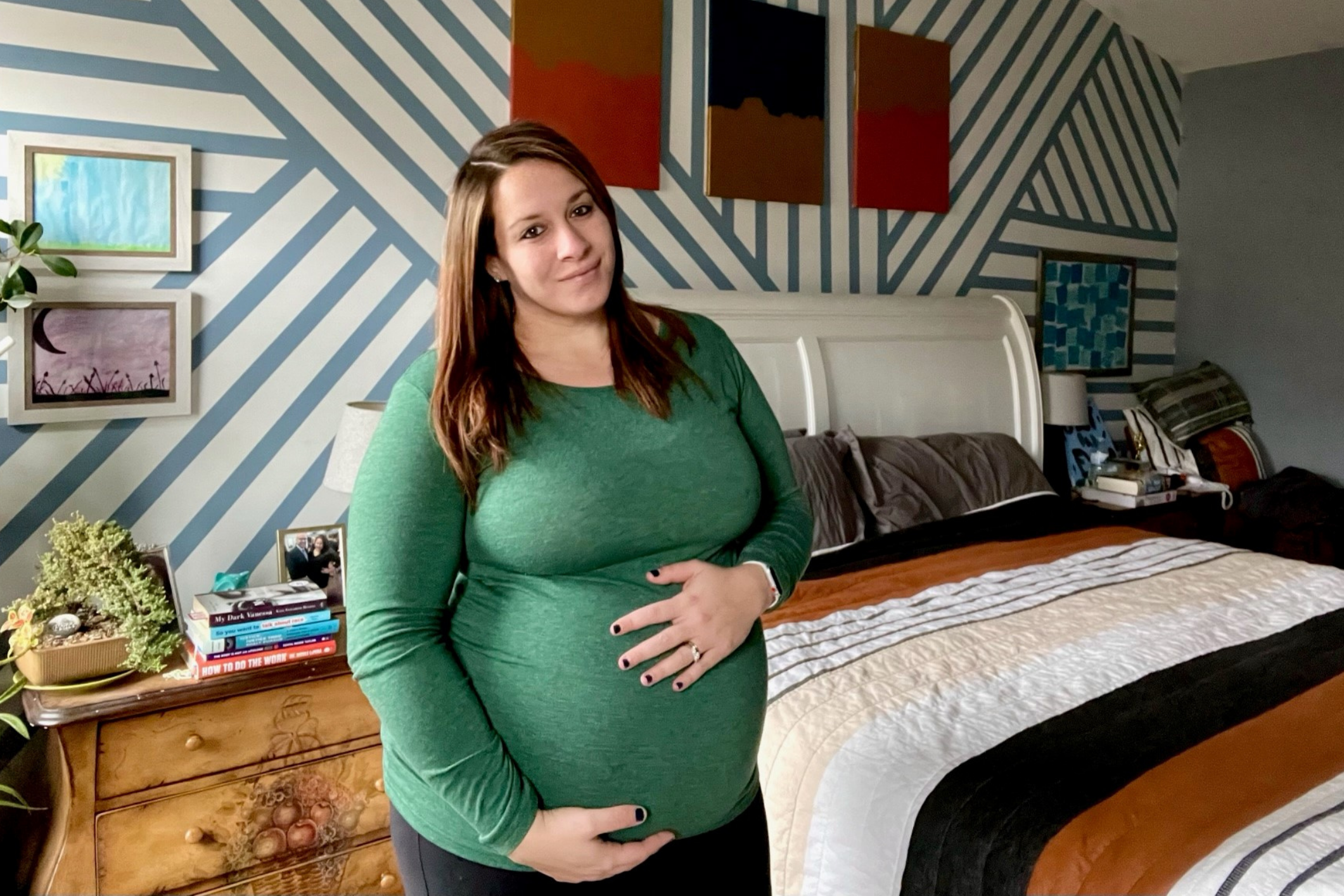 Pregnant mom cradles her belly, wearing a green long sleeve shirt, standing in a bedroom with striped walls and striped bedding.