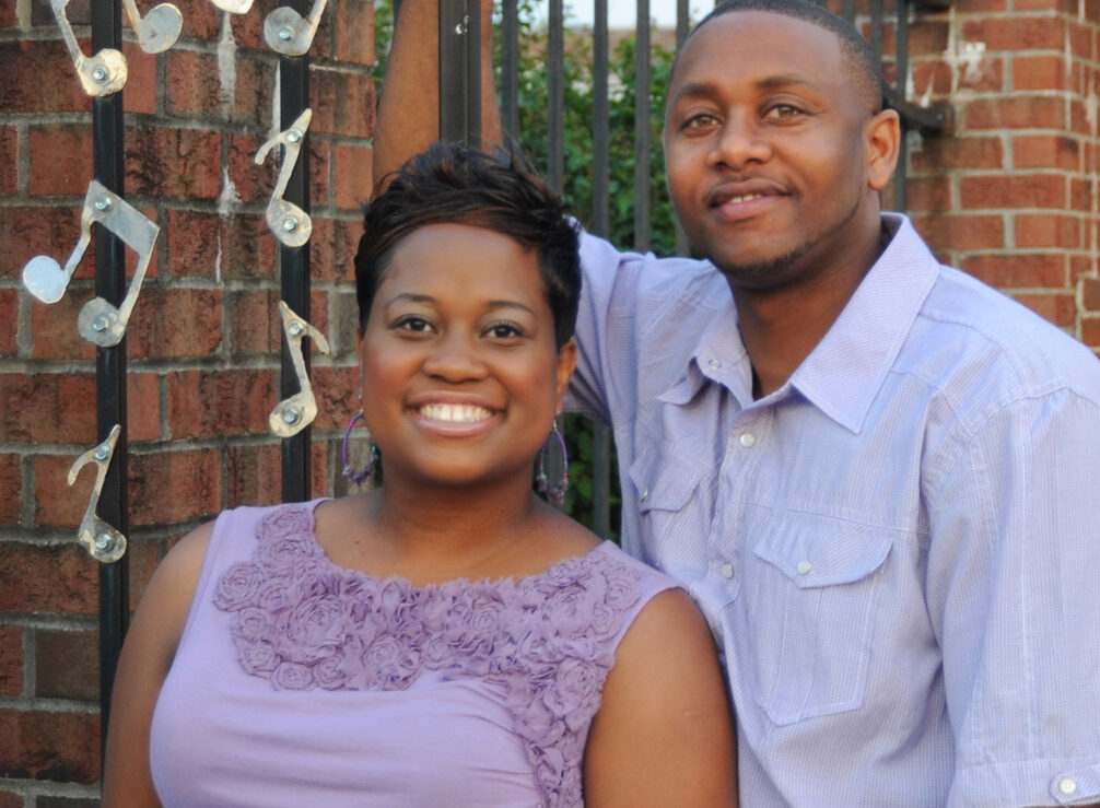 Smiling mom and dad standing in front of red brick structure, with handing musical notes.