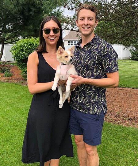 Couple smiling outside holding a small dog between them.