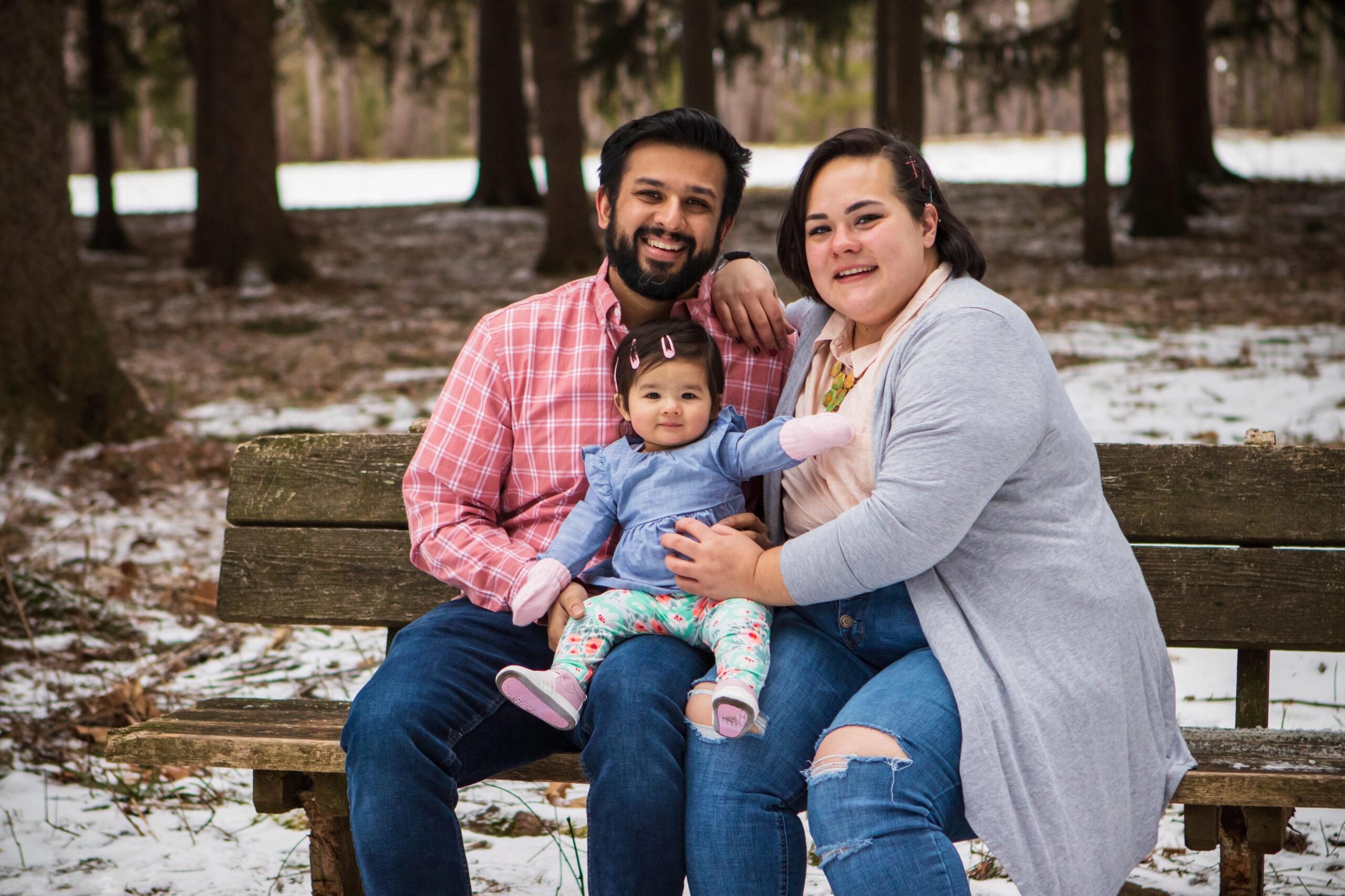 Smiling mom, dad and baby girl sitting on a bench in the snow.