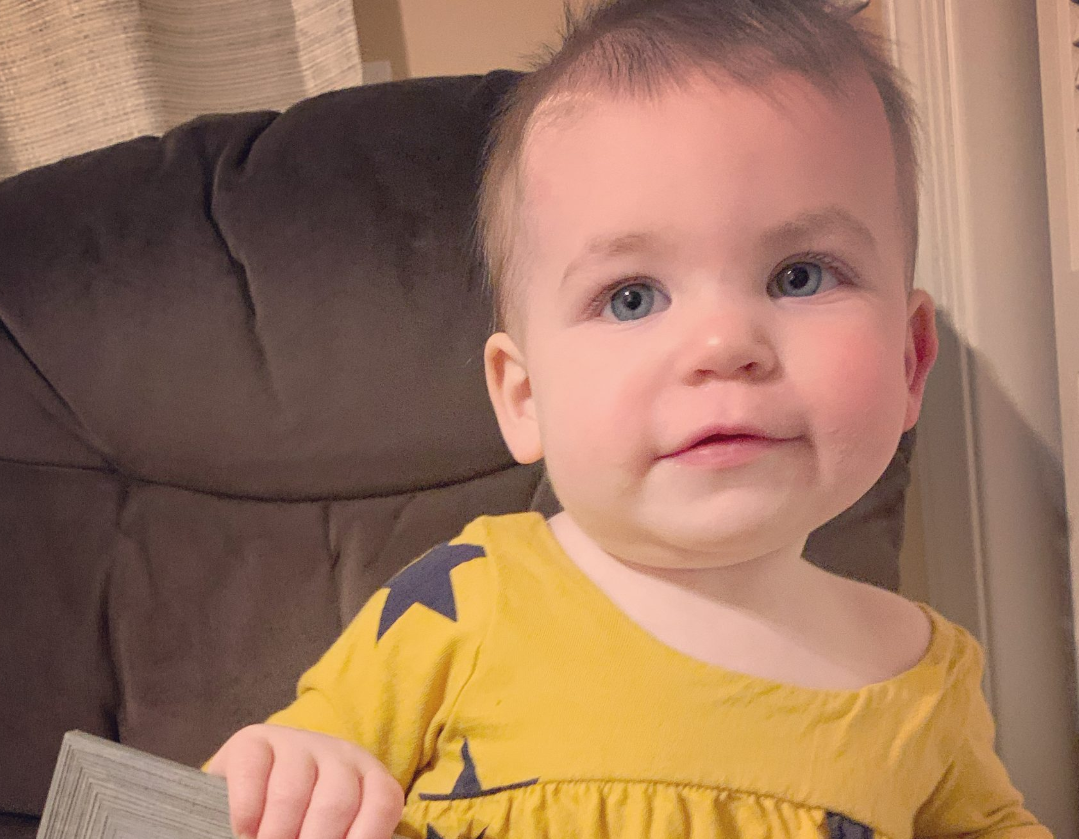 Baby girl, Genevieve, sitting in a brown chair wearing a yellow outfit with purple stars.