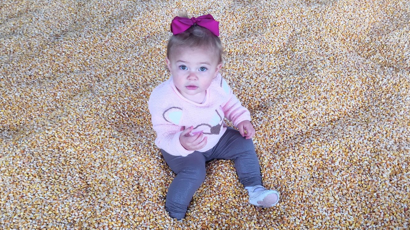 Young baby girl sitting on top of pile of corn kernels wearing a pink sweater and purple bow in her hair.