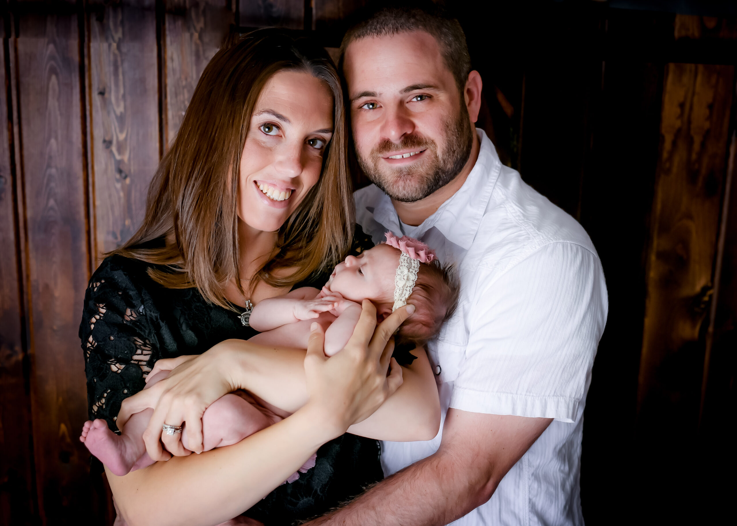 Mom and dad smiling, holding newborn baby girl with a pink and white headband.