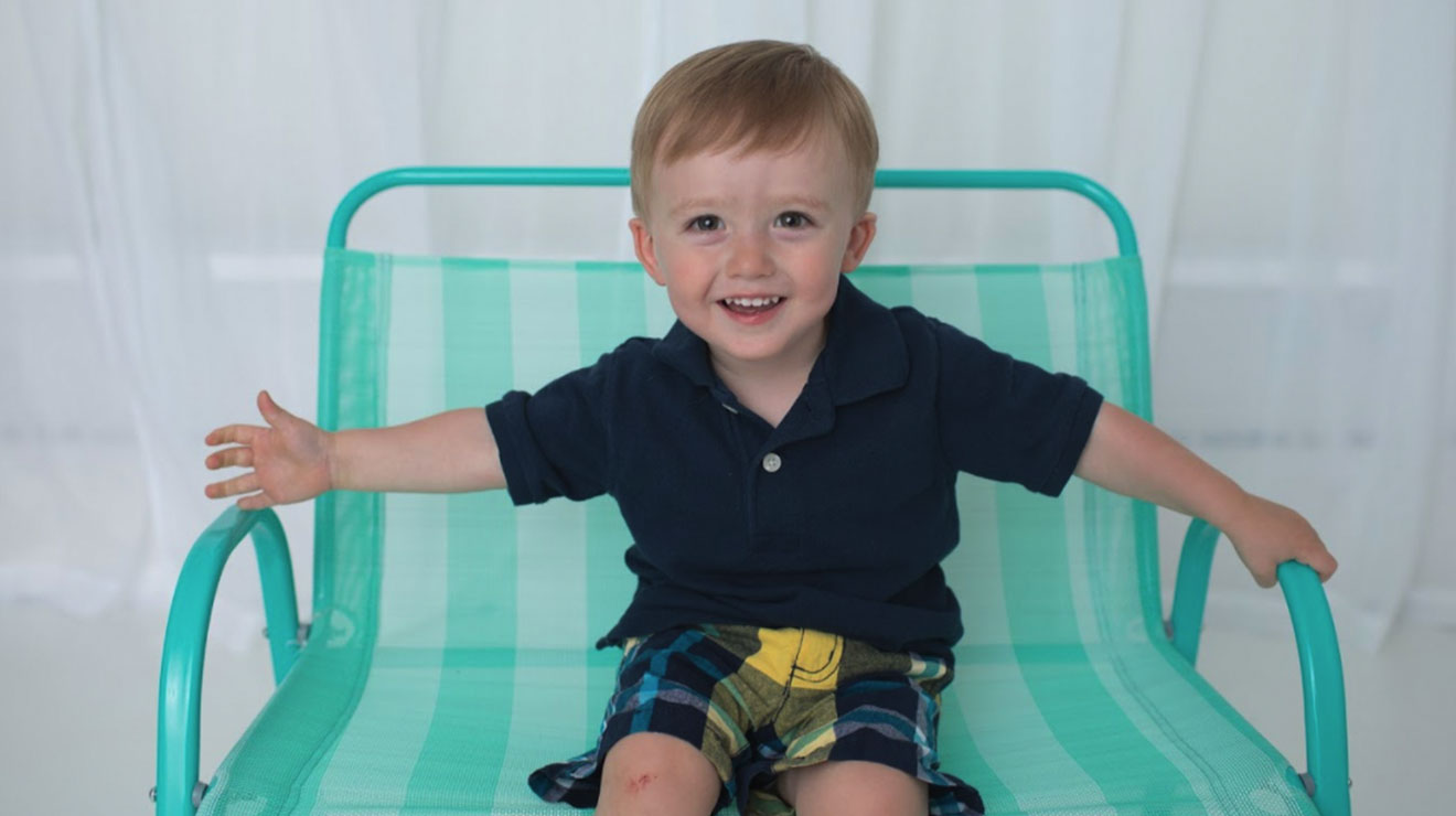 Smiling toddler boy sitting on a teal and white striped chair, wearing a navy blue polo shirt and plaid shorts.