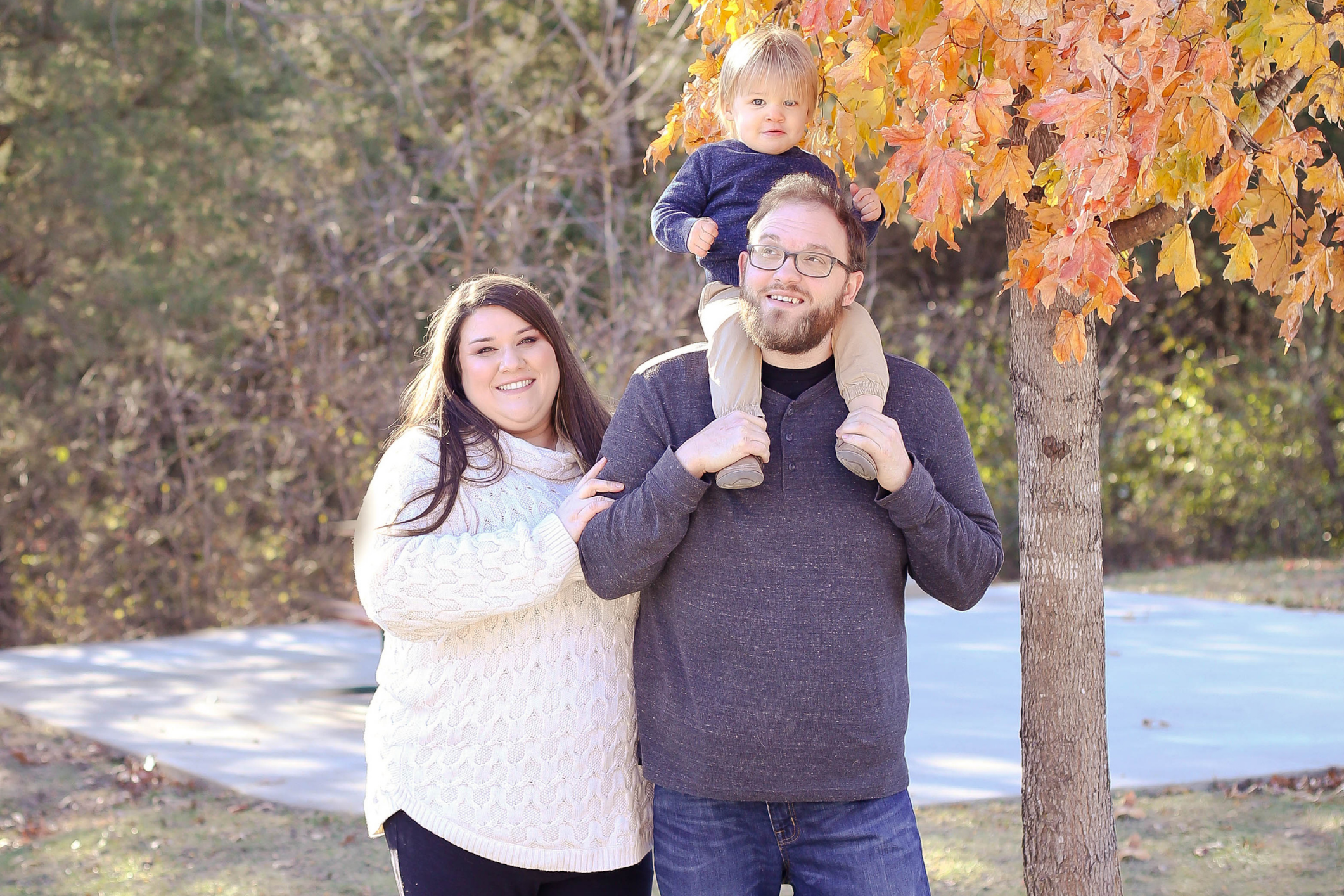 Smiling mom next to dad holding baby girl on his shoulders, all outside in the fall.