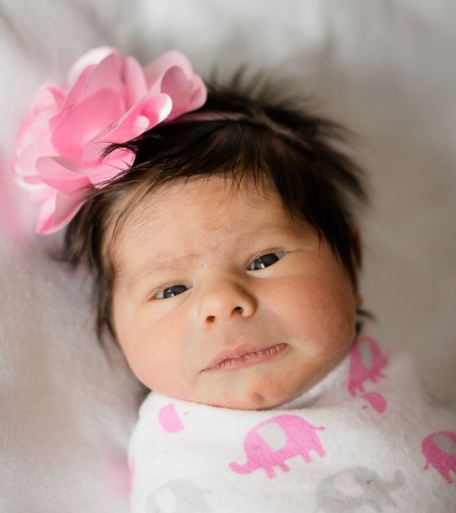 Baby girl wearing a pink, white, and gray elephant pattern swaddle, with a pink flower headband.