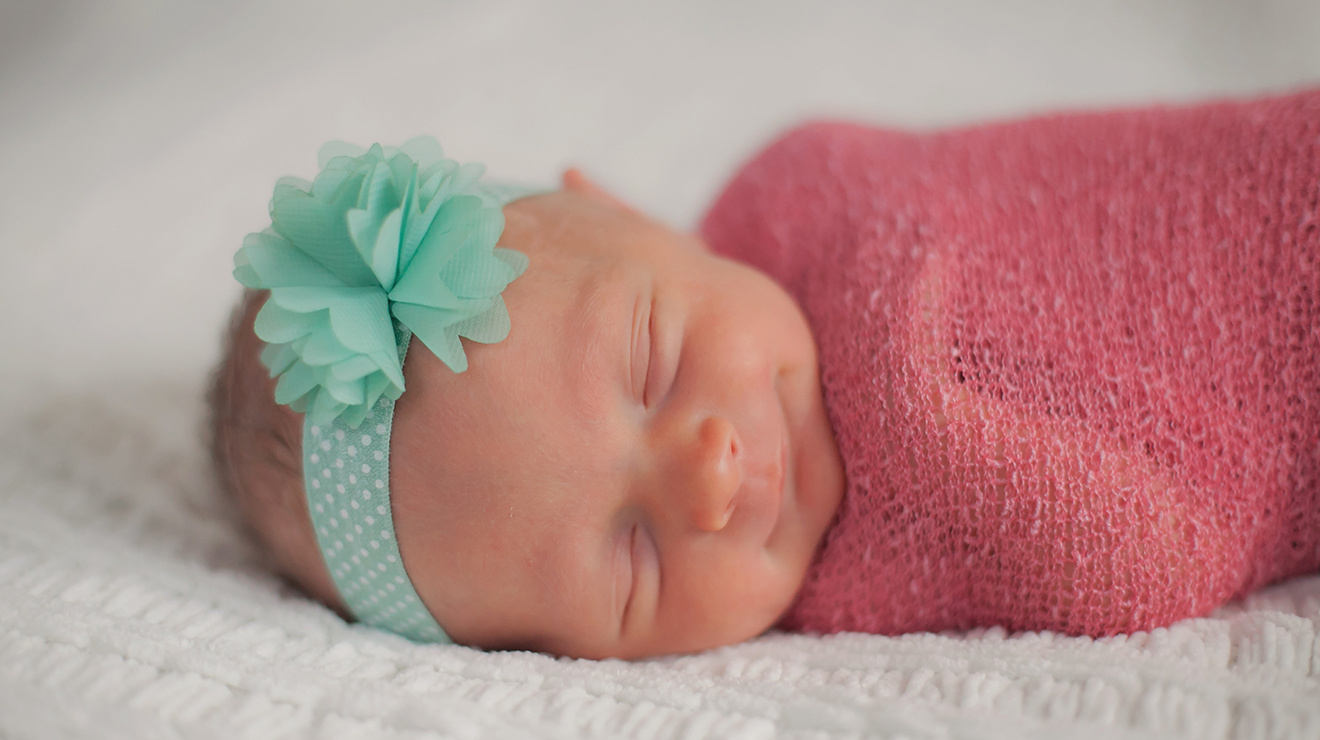 Smiling newborn baby girl laying down in a pink swaddle wearing a teal flower headband.