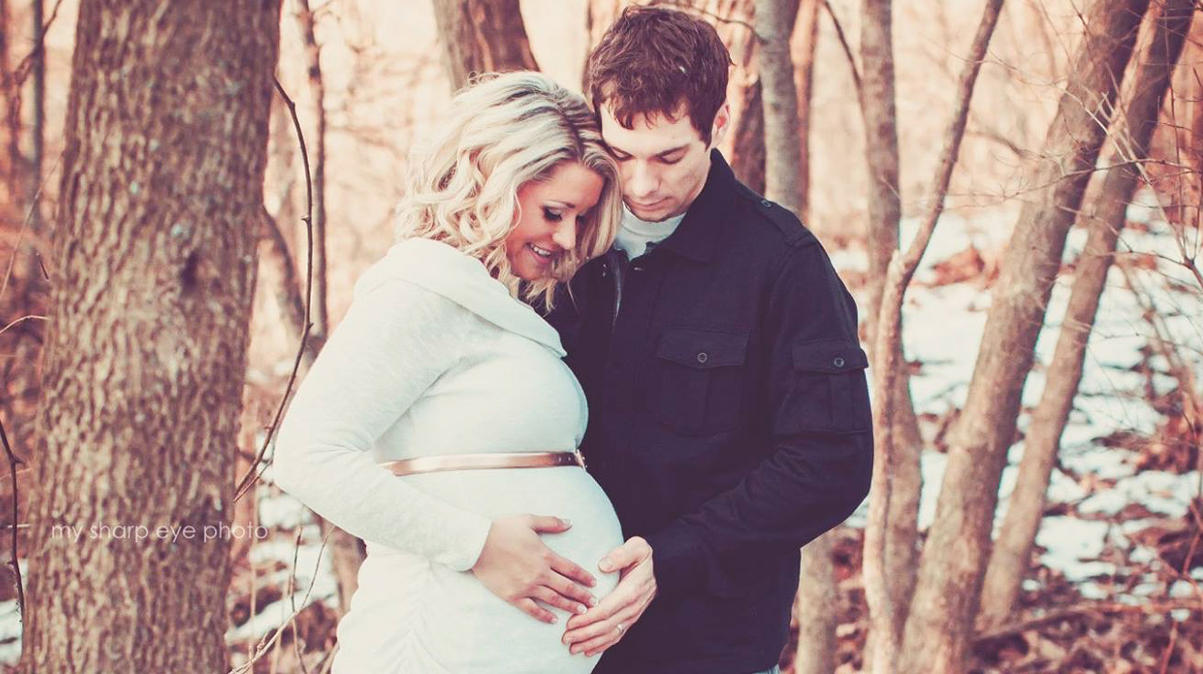Pregnant woman and her husband embracing in front of trees.