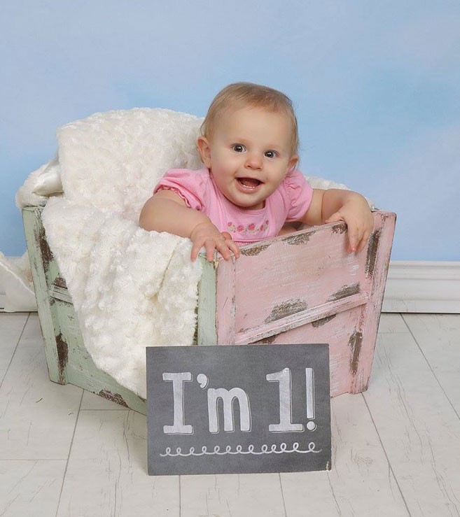 Smiling one year old girl in a pink outfit sitting in a pink and green wooden crate with a fluffy white blanket. A sign is in front of her that reads "I'm 1!"