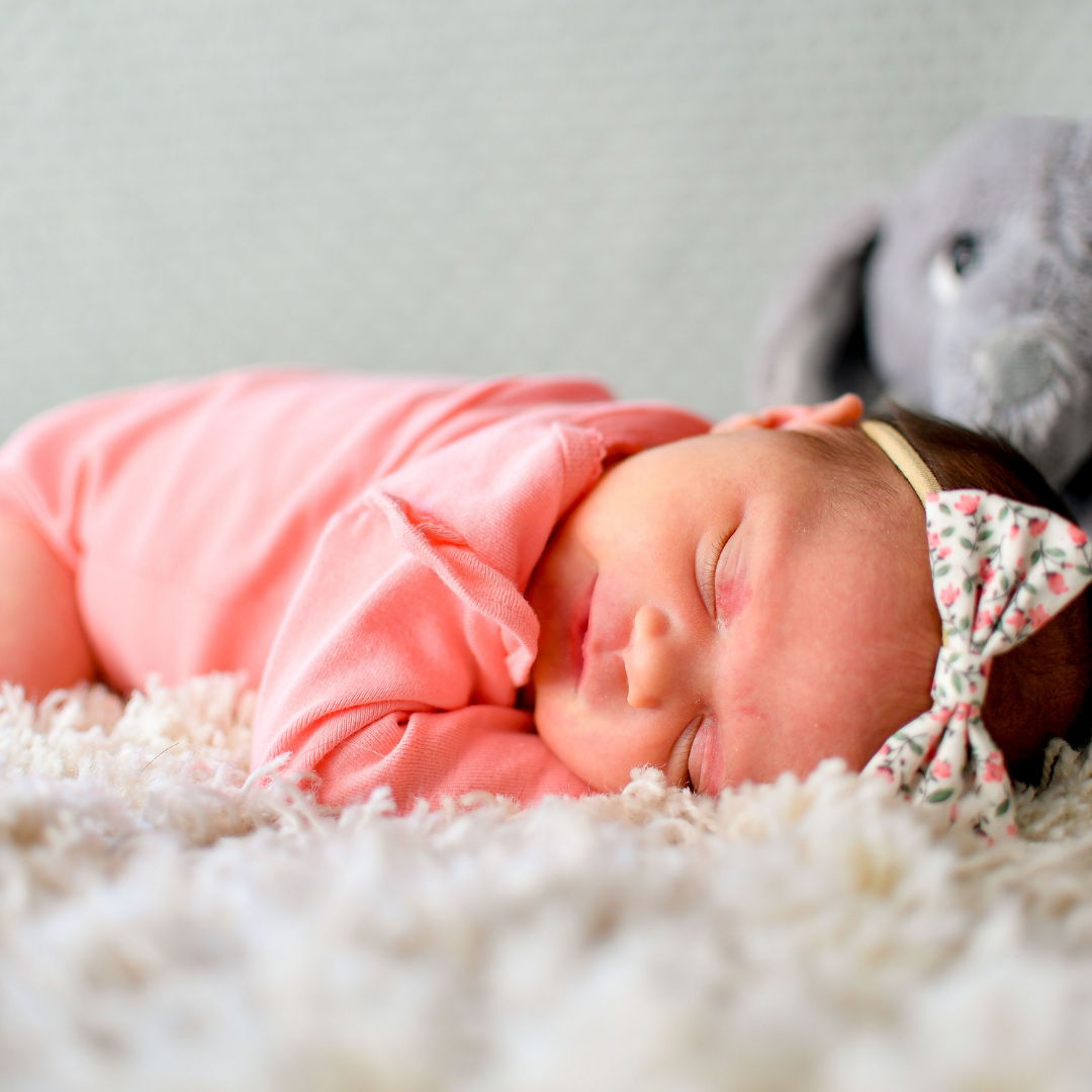 Newborn baby girl laying down in a pink onesie wearing a floral bow headband.