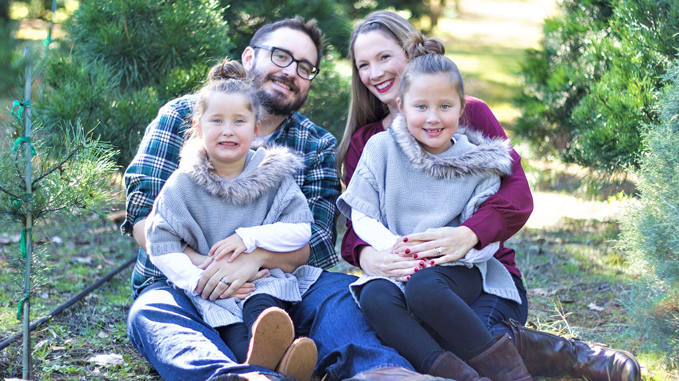 Smiling mom, dad and twin girls, all sitting on the ground near fresh trees