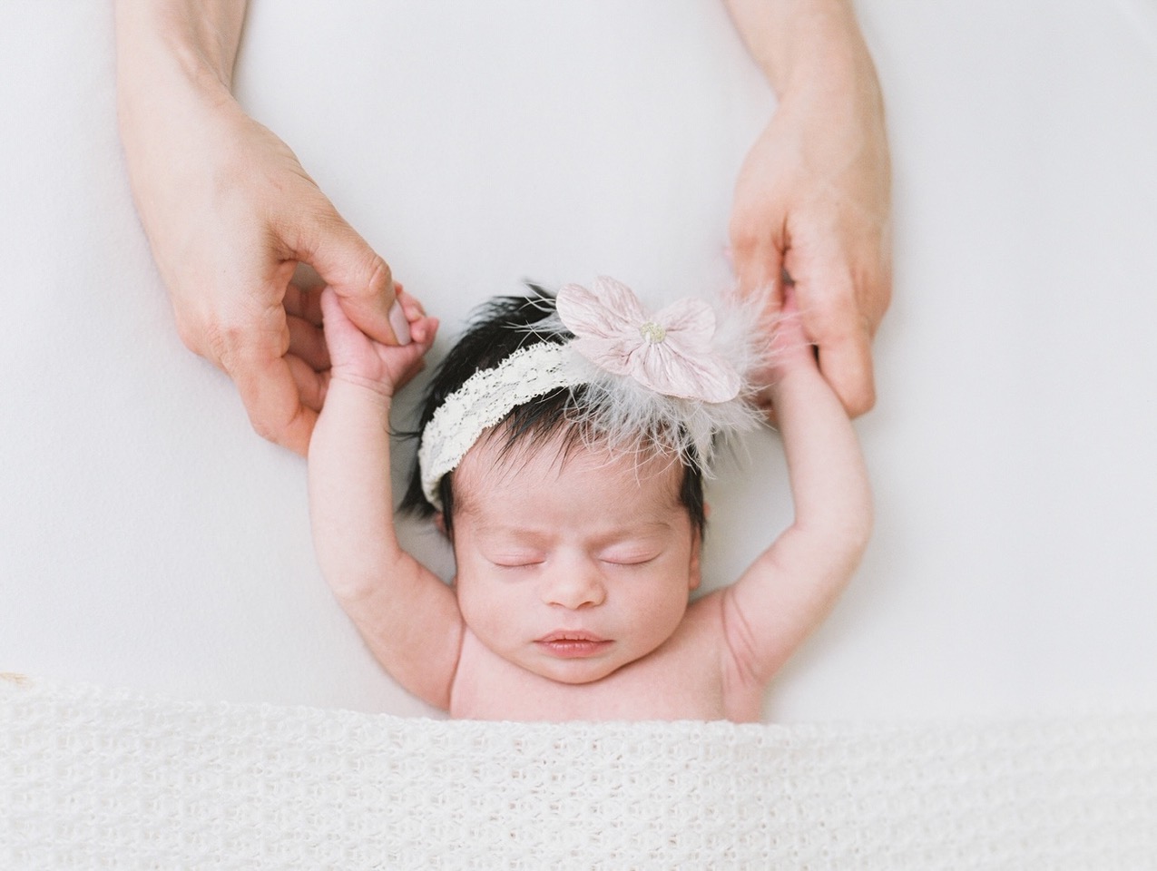 Newborn baby girl with brown her wearing a white and light pink headband with her arms above her head.