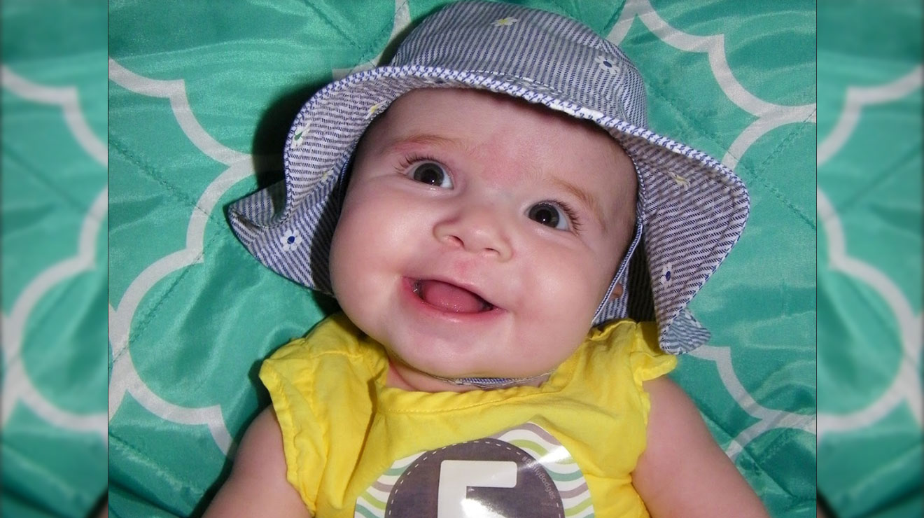 Smiling baby girl wearing a yellow outfit and purple hat, laying on a teal and white background.