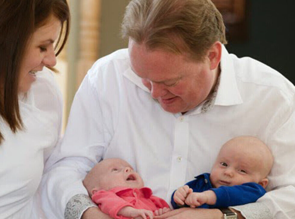 Smiling mom and dad, both in white shirts, holding boy-girl twins.