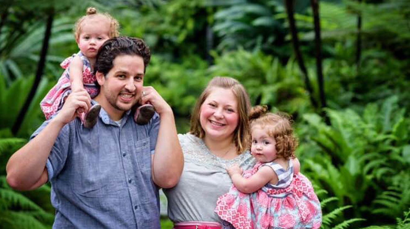 Smiling mom, dad and two twin baby girls, standing outside near trees.