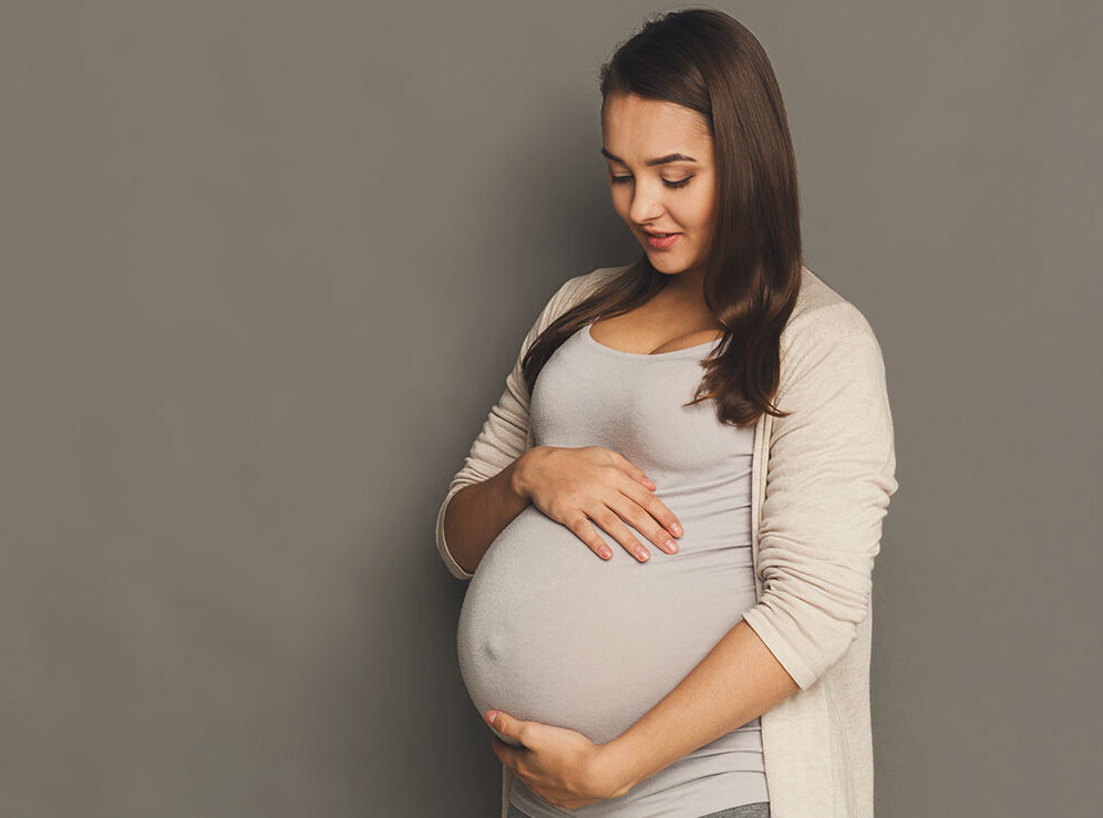 Smiling mom cradling her baby bump, wearing a tan top and sweater.