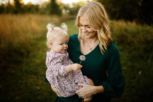 Standing in a field, mom, wearing a dark green dress, holding her baby girl, wearing a printed dress and pigtails, and holding a dandelion.