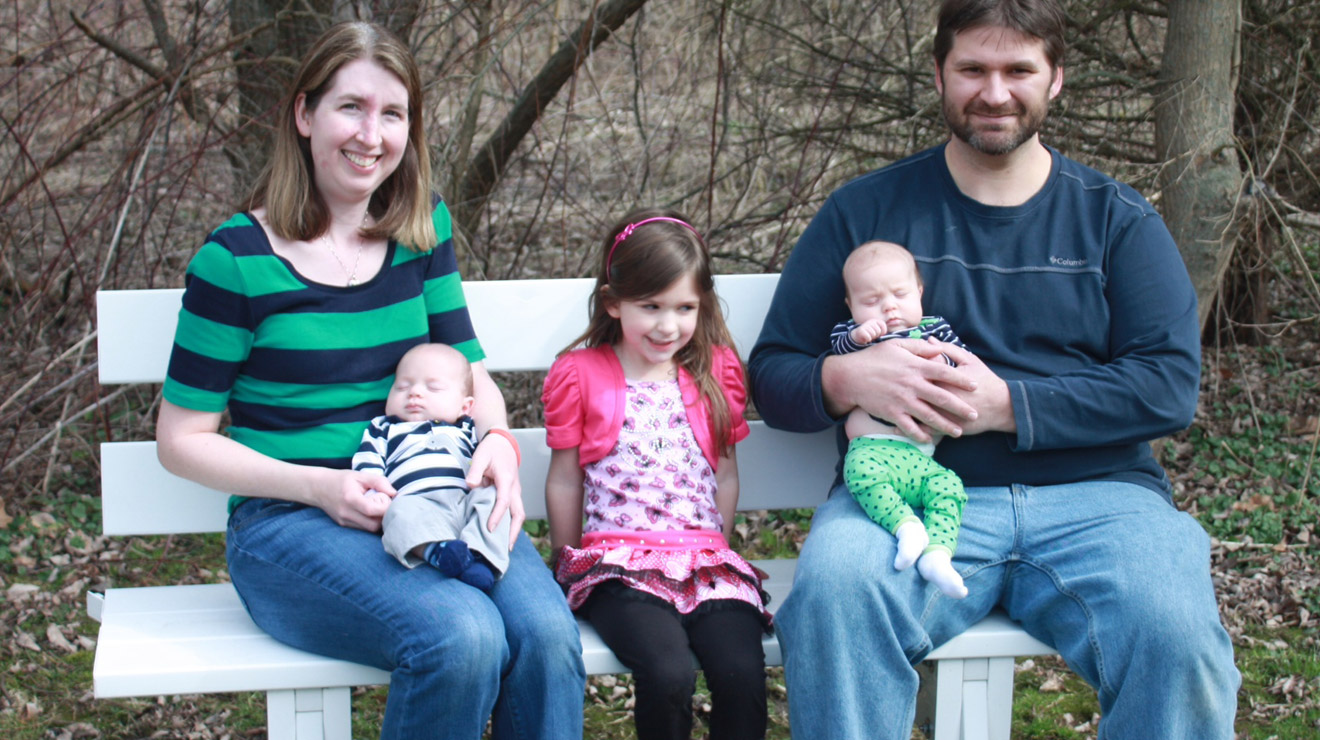 Mom and dad each holding one twin with their adolescent daughter in between them, all sitting on a bench.